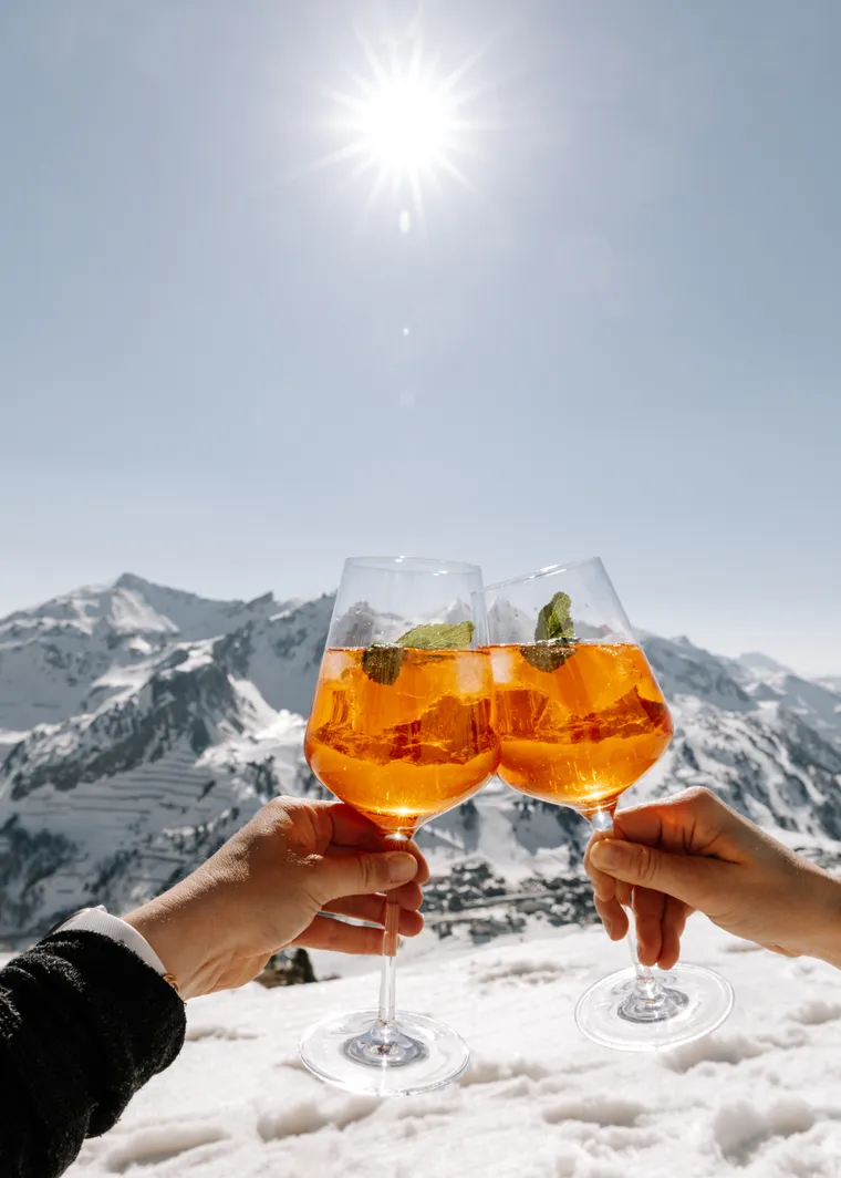 Two Aperol Spritz glasses clinking in sunny snowy Alpine mountain landscape